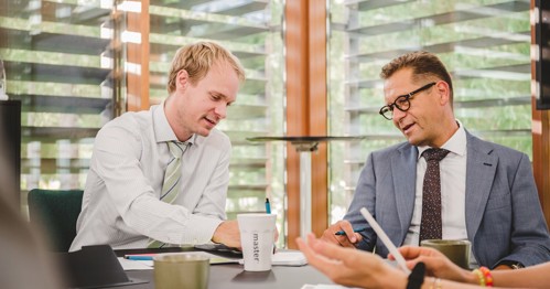 Business Meeting Discussion: Two men in business attire sitting at a table in a modern office space, engaged in a discussion while reviewing personality assessments and using electronic devices.