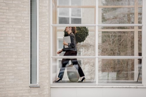 A woman in business attire walking through a modern office corridor with glass walls, carrying a laptop and looking ahead with a focused expression.