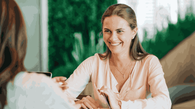 Ung kvinna med långt hår och ljus topp ler mot en kvinna som hon sitter mittemot.