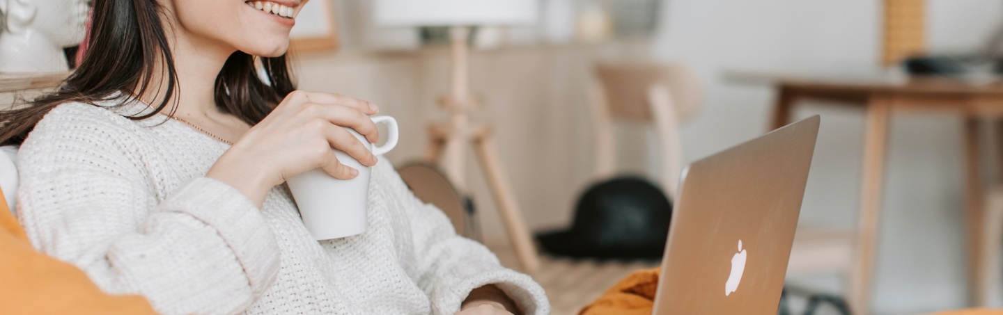 Kvinna med en kopp kaffe i handen framför sin laptop.